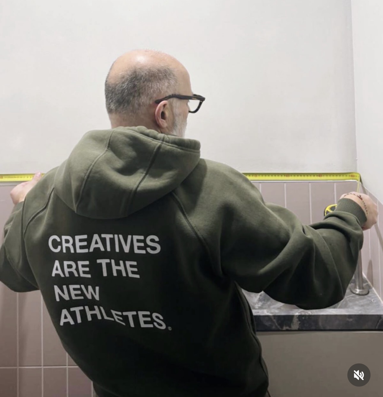 Seen from behind on a site visit, wearing a hoodie that reads ‘Creatives are the new athletes’, holding a tape measure against a tiled wall.